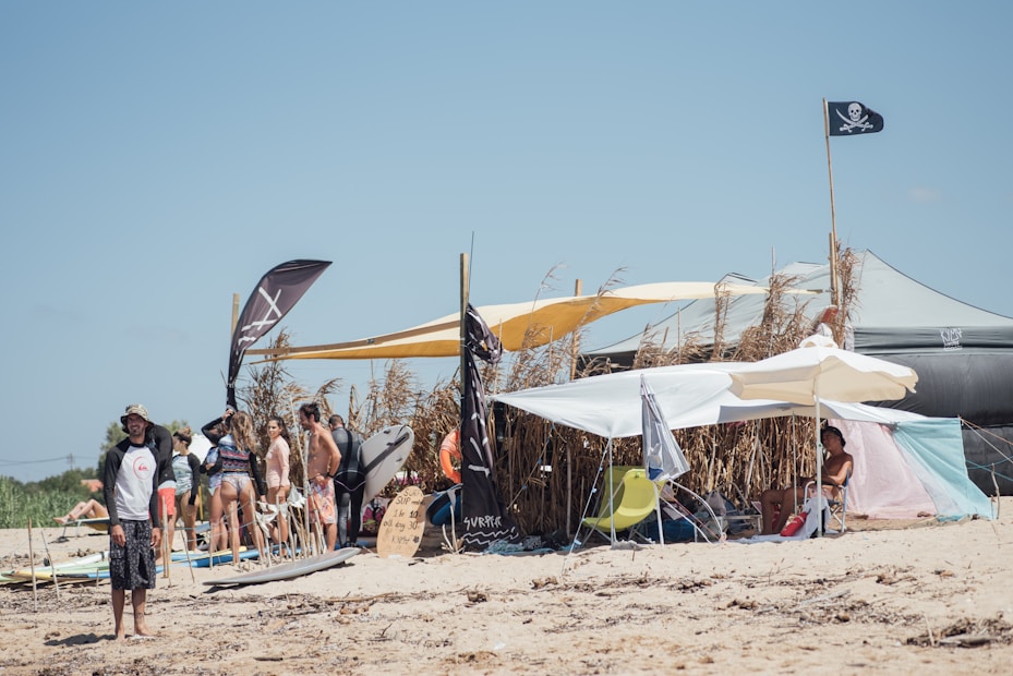 A beach scene with a group of people under makeshift tents and umbrellas. Surfboards and flags are visible, with a pirate flag prominently displayed. The background shows tall grasses and the sky is clear.