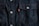 Dark blue denim fabric featuring a close-up of a front-pocket design with contrasting orange stitching, white button closures, and a small red tag with white text.
