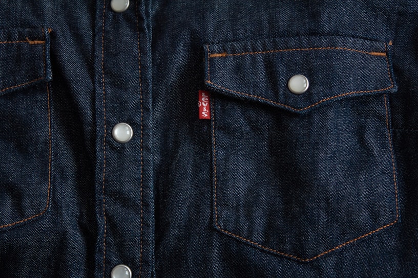 Dark blue denim fabric featuring a close-up of a front-pocket design with contrasting orange stitching, white button closures, and a small red tag with white text.
