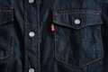 Dark blue denim fabric featuring a close-up of a front-pocket design with contrasting orange stitching, white button closures, and a small red tag with white text.