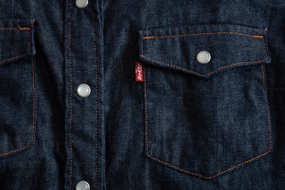 Dark blue denim fabric featuring a close-up of a front-pocket design with contrasting orange stitching, white button closures, and a small red tag with white text.