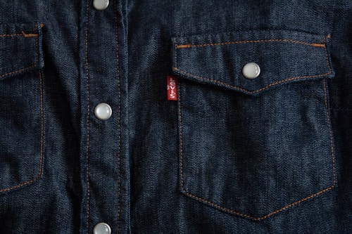 Dark blue denim fabric featuring a close-up of a front-pocket design with contrasting orange stitching, white button closures, and a small red tag with white text.