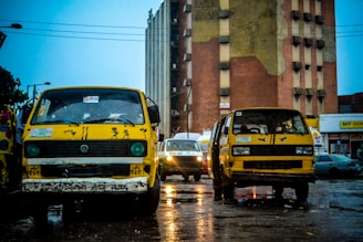 Two yellow buses are parked on a wet street in front of a tall, aged building. The area appears to have just experienced rain, as the pavement is shining with reflections. Other vehicles are visible in the background, and some colorful clothing is seen on the left.
