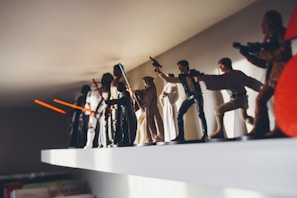 A collection of Star Wars action figures displayed on a white shelf, featuring characters like Jedi with lightsabers and other iconic figures in dynamic poses.