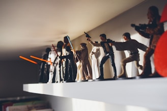 A collection of Star Wars action figures displayed on a white shelf, featuring characters like Jedi with lightsabers and other iconic figures in dynamic poses.
