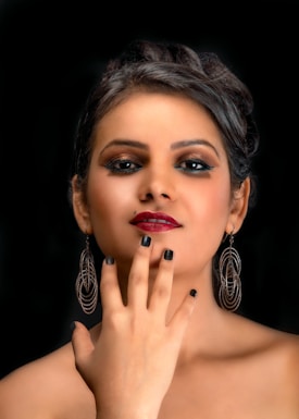 A woman with dark makeup and vibrant, glossy lipstick poses against a black background. Her hair is styled elegantly, and she is wearing large, intricate hoop earrings. Her hand, with nails painted in a deep black shade, is placed gracefully near her lips, adding a touch of sophistication.