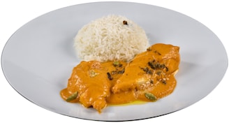 A dish featuring a serving of white rice paired with a portion of chicken covered in an orange, creamy sauce. The sauce is garnished with herbs and spices, possibly including cardamom pods and cloves. The presentation is on a simple white plate.