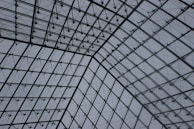 An architectural structure with a gridded and latticed pattern made of metal and glass. The design features intersecting beams and cables creating a geometric framework that curves upward. The background of the structure shows a cloudy, grey sky.
