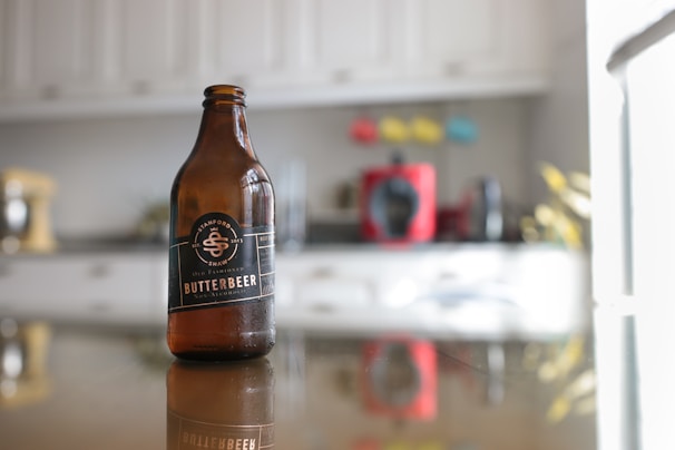 A brown glass bottle labeled 'Butterbeer' is positioned on a reflective surface in a kitchen setting. The background shows blurry kitchen appliances and cabinetry, with several colored decorations adding contrast.