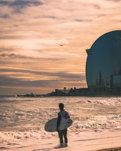 A surfer stands on the sandy beach, looking out to sea with a surfboard in hand. The ocean waves gently roll towards the shore. In the distance, a large modern building with a curved design rises against a cloudy sky, and a plane can be seen flying overhead.