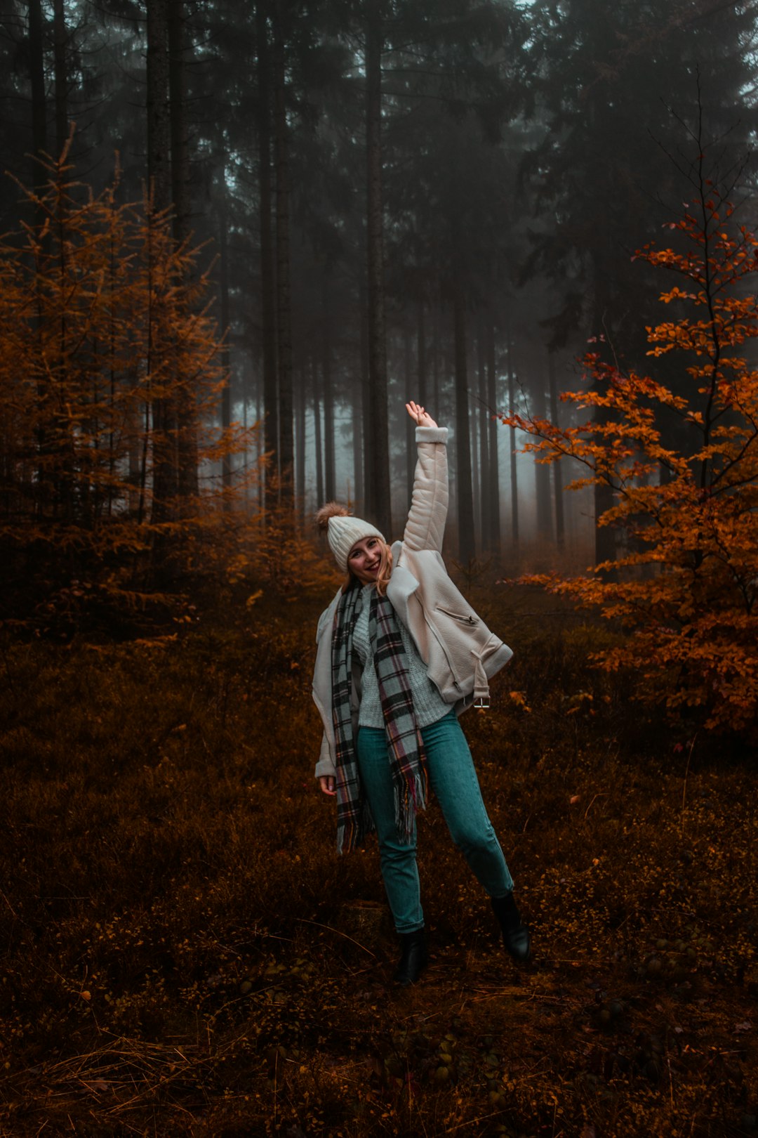 smiling woman wearing white jacket and hat standing while raising left hand surrounded with green trees in foggy day