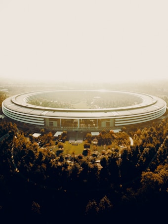 Aerial view of a large, circular building surrounded by trees. The structure has a sleek, modern design with a reflective surface and appears to be part of a sprawling corporate campus. Sunlight filters through the haze, creating a warm, golden hue over the scene.