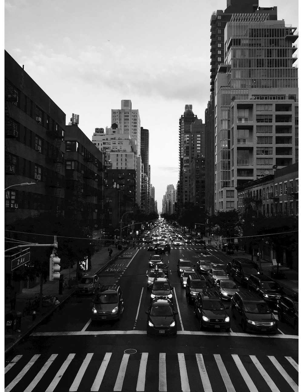 grayscale photography of people walking on pathway near buildings and different vehicles on road