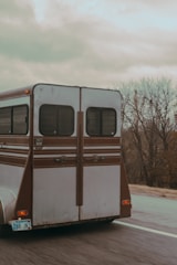 A brown and white horse trailer is being driven on a road with a Kansas license plate. The background features leafless trees and a cloudy sky, suggesting an autumn or winter setting.