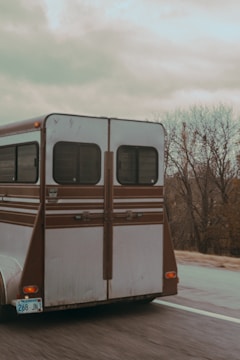 A brown and white horse trailer is being driven on a road with a Kansas license plate. The background features leafless trees and a cloudy sky, suggesting an autumn or winter setting.