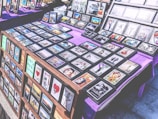 A marketplace stall features an array of framed images and posters with various designs, arranged on tables covered with purple cloth. The collection includes diverse themes such as landscapes, inspirational quotes, pop culture references, and abstract art. The frames are neatly organized, and some larger prints are displayed upright in the background.
