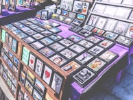 A marketplace stall features an array of framed images and posters with various designs, arranged on tables covered with purple cloth. The collection includes diverse themes such as landscapes, inspirational quotes, pop culture references, and abstract art. The frames are neatly organized, and some larger prints are displayed upright in the background.