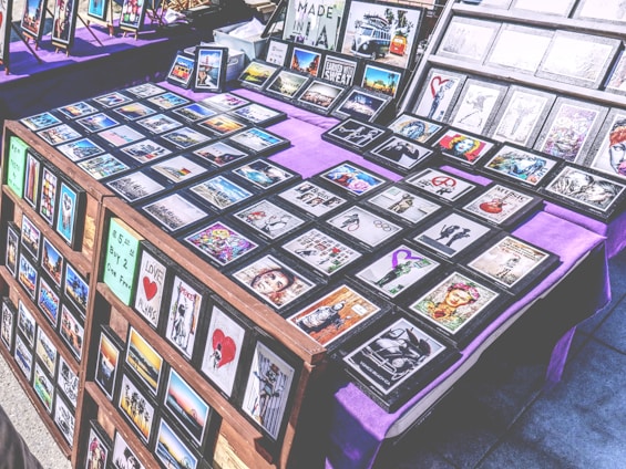 A marketplace stall features an array of framed images and posters with various designs, arranged on tables covered with purple cloth. The collection includes diverse themes such as landscapes, inspirational quotes, pop culture references, and abstract art. The frames are neatly organized, and some larger prints are displayed upright in the background.