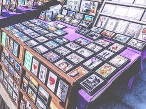 A marketplace stall features an array of framed images and posters with various designs, arranged on tables covered with purple cloth. The collection includes diverse themes such as landscapes, inspirational quotes, pop culture references, and abstract art. The frames are neatly organized, and some larger prints are displayed upright in the background.