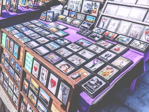 A marketplace stall features an array of framed images and posters with various designs, arranged on tables covered with purple cloth. The collection includes diverse themes such as landscapes, inspirational quotes, pop culture references, and abstract art. The frames are neatly organized, and some larger prints are displayed upright in the background.