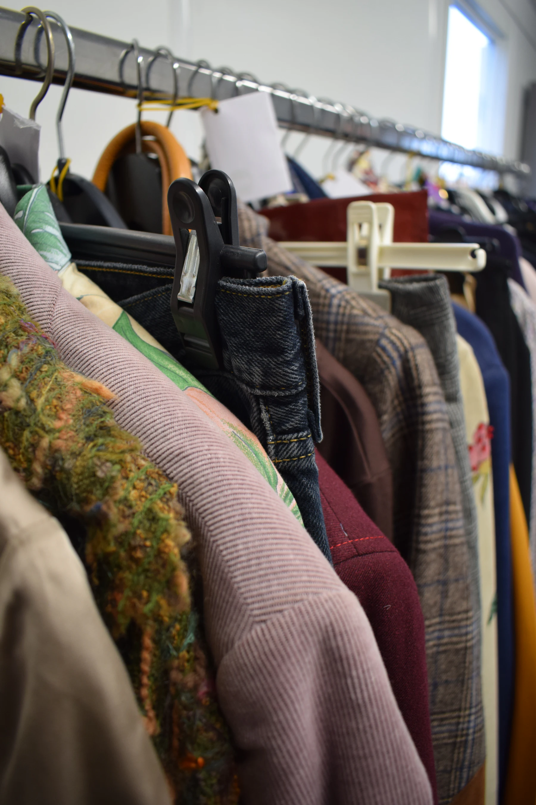 Close view along a secondhand rack: a corduroy shoulder, a plaid wool jacket, tweed and a pair of jeans on a clip hanger