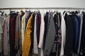 A clothing rack filled with a variety of garments, including shirts, jackets, and blazers in different colors and patterns. The assortment displays a diverse selection of fabrics and designs, indicating a collection of formal and casual wear.
