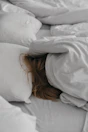 A person with long hair is seen resting beneath a light-colored duvet, surrounded by several pillows. The setting suggests a cozy and peaceful bedroom ambiance.
