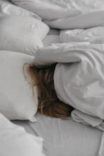 A person with long hair is seen resting beneath a light-colored duvet, surrounded by several pillows. The setting suggests a cozy and peaceful bedroom ambiance.