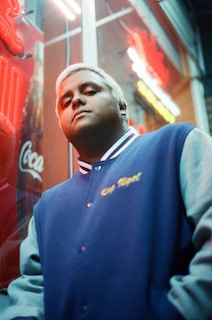 A person with short, blonde hair is wearing a blue varsity jacket with white sleeves and looking confidently at the camera. The background features neon lights and a glass window displaying the word 'Coca-Cola'.