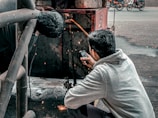 Two individuals are engaged in welding work. One person is actively welding, creating visible sparks, while the other appears to be holding a mobile phone, possibly to document or assist with the task. The setting is outdoors, with a metal structure and a street in the background, where motorcycles are visible.