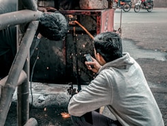 Two individuals are engaged in welding work. One person is actively welding, creating visible sparks, while the other appears to be holding a mobile phone, possibly to document or assist with the task. The setting is outdoors, with a metal structure and a street in the background, where motorcycles are visible.