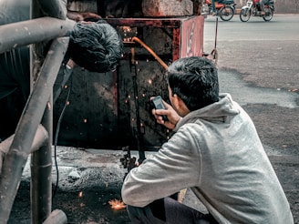Two individuals are engaged in welding work. One person is actively welding, creating visible sparks, while the other appears to be holding a mobile phone, possibly to document or assist with the task. The setting is outdoors, with a metal structure and a street in the background, where motorcycles are visible.