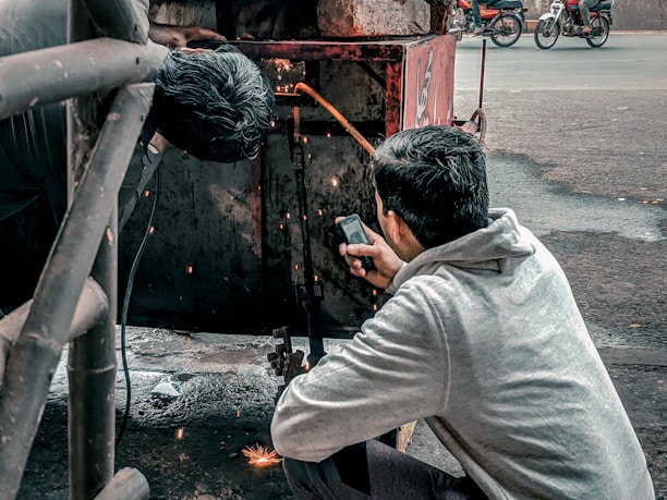 Two individuals are engaged in welding work. One person is actively welding, creating visible sparks, while the other appears to be holding a mobile phone, possibly to document or assist with the task. The setting is outdoors, with a metal structure and a street in the background, where motorcycles are visible.