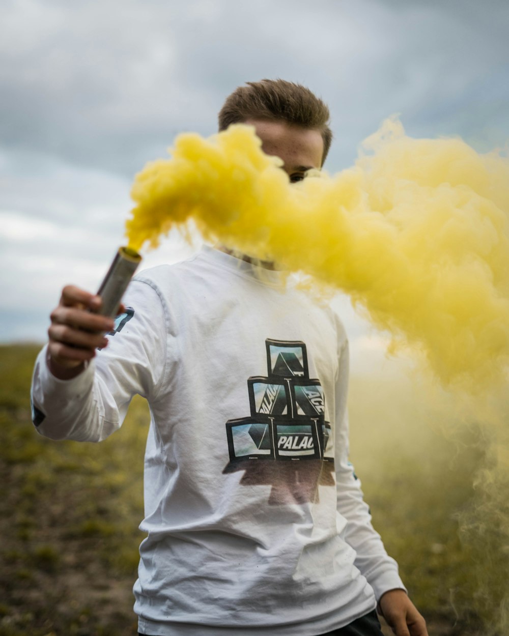 man wearing white, black, and green printed crew-neck long-sleeved shirt standing while holding yellow smoke on green field
