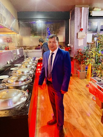 A man in a blue suit stands beside a buffet line with multiple covered dishes. The environment appears to be a restaurant with wooden floors, decorative plants, and warm lighting.