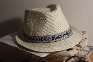 A woven straw hat with a geometric patterned black and white band around the base rests on top of a pile of papers and a woven basket. The hat's surface texture is detailed and natural, casting a soft shadow on a light wall in the background.
