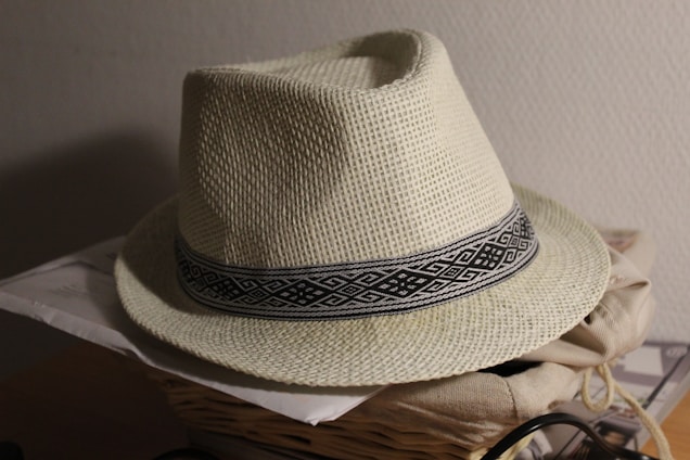 A woven straw hat with a geometric patterned black and white band around the base rests on top of a pile of papers and a woven basket. The hat's surface texture is detailed and natural, casting a soft shadow on a light wall in the background.