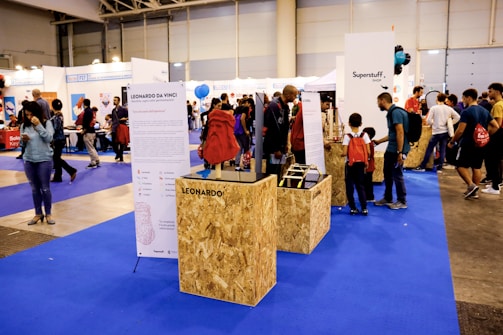 The scene takes place in a large indoor exhibition hall with various booths and displays. People of different ages, including children and adults, are walking around, observing exhibits, and engaging with the displays. A focal point is a booth featuring information about Leonardo da Vinci, with text and images, and it appears to be drawing interest from several visitors. There are also other booths in view with different exhibits and displays. The floor is covered with blue carpet, and several banners and signs can be seen around.