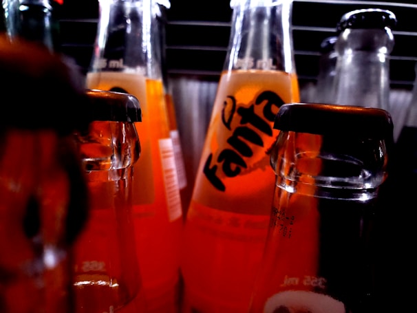 Several glass soda bottles with orange liquid are arranged closely together. The central bottle is prominently labeled Fanta. Light reflects off the glass, creating a vivid contrast against a darker background.
