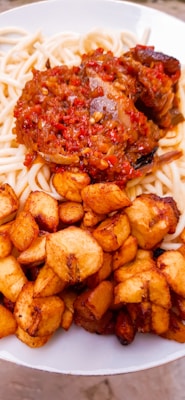 A plate of pasta topped with a rich, red tomato-based sauce containing vegetables and possibly meat. Accompanying the pasta and sauce are golden brown fried plantains arranged neatly on the side.