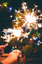 Sparklers held in hand are glowing brightly with a backdrop of colorful, blurred festive lights. The scene suggests a celebratory atmosphere.