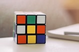 A Rubik's Cube sits on a white surface, partially solved with various bright colors on its visible faces. There is a blurred background and the lighting suggests a soft, natural light source.