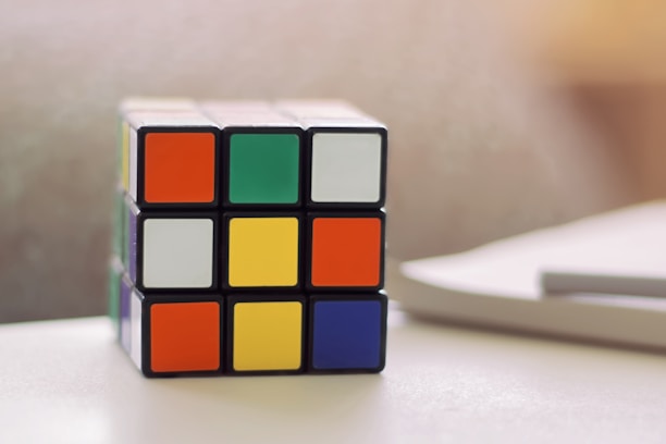 A Rubik's Cube sits on a white surface, partially solved with various bright colors on its visible faces. There is a blurred background and the lighting suggests a soft, natural light source.