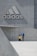 Two people are standing near a large concrete structure with a prominent Adidas logo on the wall. One person appears to be an adult wearing a blue shirt, bending down towards a child in a yellow shirt. The setting appears urban and minimalist, with a gray concrete background.