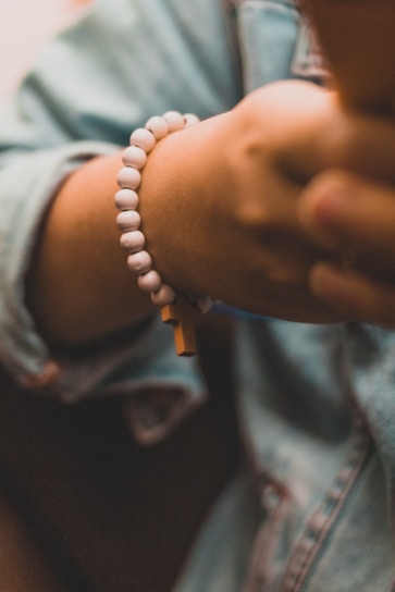 A hand wearing a white beaded bracelet, with one bead shaped like a cross. The hand is partially shown against a blurred background, and the person appears to be wearing a denim jacket.