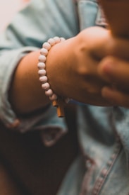 A hand wearing a white beaded bracelet, with one bead shaped like a cross. The hand is partially shown against a blurred background, and the person appears to be wearing a denim jacket.