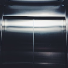 Smooth metallic elevator doors with a reflective surface, catching light at the top and bottom edges. The doors are centered and closed, with a dark seam running down the middle.