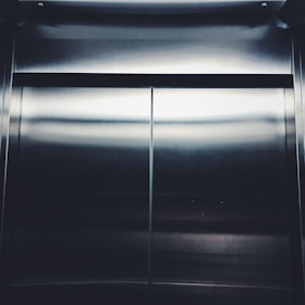 Smooth metallic elevator doors with a reflective surface, catching light at the top and bottom edges. The doors are centered and closed, with a dark seam running down the middle.