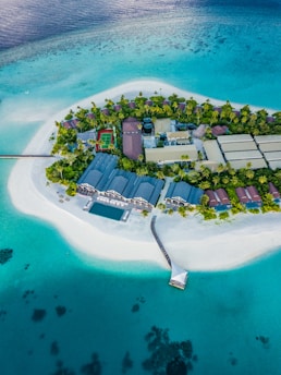 An aerial view of a tropical island resort surrounded by turquoise waters. The island features lush greenery, a series of buildings with red and beige roofs, and a central courtyard area. There is a tennis court and a long wooden pier extending into the ocean. The white sandy beach contrasts with the vibrant blue hues of the surrounding ocean.
