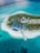 An aerial view of a tropical island resort surrounded by turquoise waters. The island features lush greenery, a series of buildings with red and beige roofs, and a central courtyard area. There is a tennis court and a long wooden pier extending into the ocean. The white sandy beach contrasts with the vibrant blue hues of the surrounding ocean.